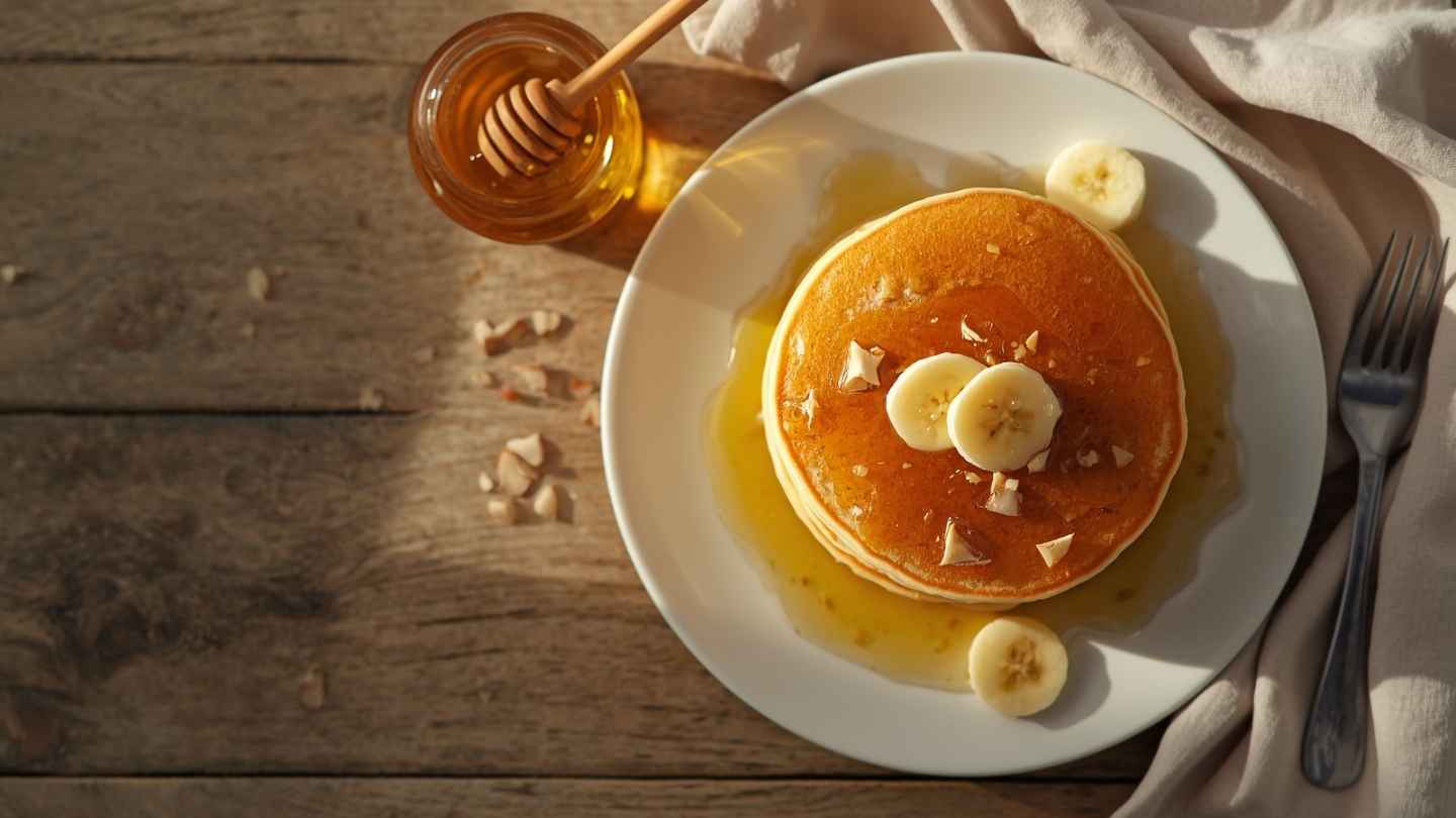 Pancake madu tanpa gula tambahan yang lembut dengan topping pisang dan madu asli di atas piring putih