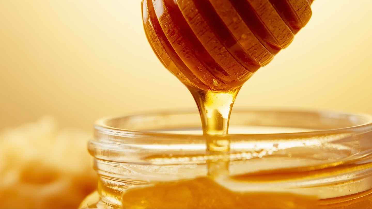 Madu murni menetes dari sendok kayu ke dalam toples kaca dengan sarang lebah di latar belakang, menggambarkan kandungan protein dan enzim alami dalam madu