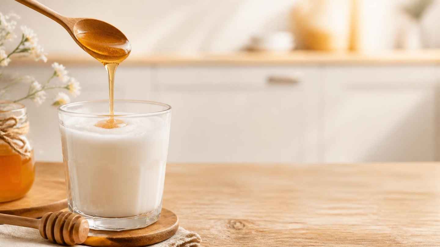 Segelas susu hangat dengan madu menetes dari sendok di atas meja kayu, manfaat susu dan madu untuk kesehatan